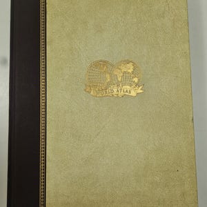 May include: A vintage world atlas with a gold emblem and the words "WORLD ATLAS" on the cover. The cover is a light tan color with a dark brown spine. The spine has a gold decorative border.