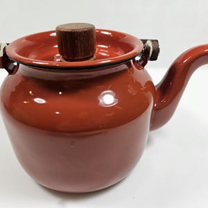 May include: A reddish-brown ceramic teapot with a wooden lid and handle accents. The teapot has a rounded body, a curved spout, and two small handles on either side. The lid is cylindrical and made of wood.