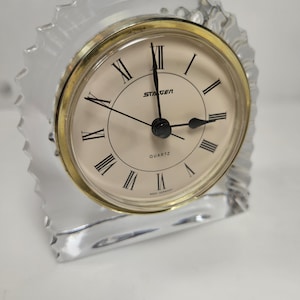 May include: A clear glass quartz clock with a gold-colored bezel and a cream-colored face. The clock has black Roman numerals and black hands. The clock is marked "STAGER QUARTZ" and "WEST GERMANY". The clock is in a clear glass frame.