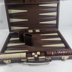 May include: Open backgammon set with a dark brown exterior and a light brown playing surface. The set includes dice, checkers, and dice cups. The case is open, revealing the game pieces and playing board.