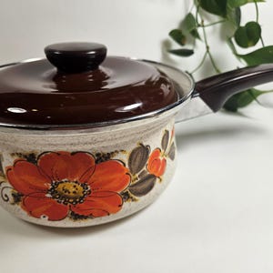 May include: A brown enameled saucepan with a floral design. The saucepan has a brown handle and a lid with a brown knob. The floral design features orange, brown, and white flowers.