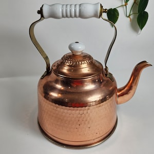 May include: A copper kettle with a hammered finish and a white porcelain handle and lid. The kettle has a brass spout and handle.