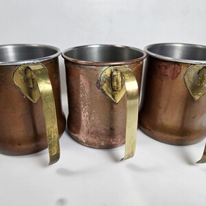 May include: Three copper mugs with brass handles. The mugs have a cylindrical shape with a silver-colored interior rim. The handles are attached with a decorative brass plate. The copper has a warm, aged patina.