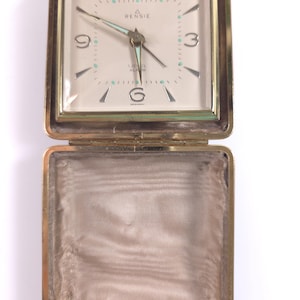 May include: A gold-tone metal travel alarm clock with a square case that opens to reveal a cream-colored face. The clock features the text RENSIE, 17 JEWEL ALARM, and GERMANY. The dial has silver-tone numerals, light blue accents, and hands.
