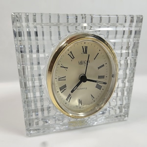 May include: A clear, square crystal clock with a gold-tone face and Roman numerals. The clock face reads "MIKASA QUARTZ". The crystal has a textured, grid-like pattern. A decorative desk clock.