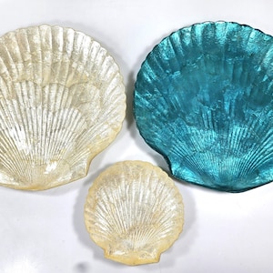 May include: Three decorative seashell-shaped dishes. Two are off-white, and one is a vibrant teal. The shells vary in size, with the largest measuring approximately 12 inches in diameter. The shells have a textured, iridescent finish.
