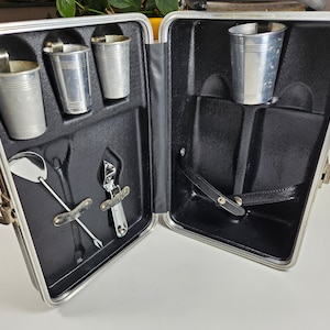 May include: A silver metal travel bar set with four cups, a bottle opener, and a cocktail spoon. The set is in a silver metal case with a black interior.