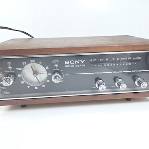 May include: A rectangular Sony Solid State AM radio with a wooden casing and a black front panel. It features an analog clock face on the left, a horizontal AM frequency tuning scale, and five silver control knobs for tone, volume, and tuning.