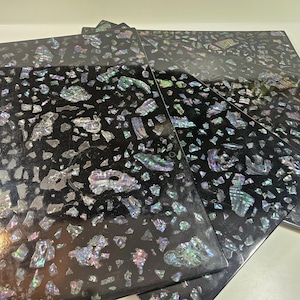 May include: A collection of black square coasters with a mosaic-like design. The coasters are covered in iridescent, multi-colored pieces that reflect light. The overall effect is a dark, textured surface with shimmering accents.