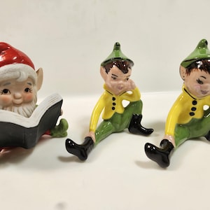 May include: Three ceramic elf figurines. One elf wears a red hat and reads a book. The other two elves wear green hats, yellow jackets, green pants, and black boots. The figurines are sitting down.