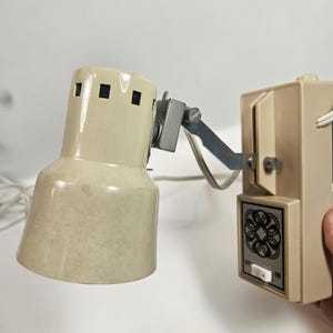 May include: A vintage beige wall-mounted lamp with a rectangular base and adjustable arm. The lamp shade has a cylindrical shape with square cutouts. The base features an on/off switch and decorative detailing.