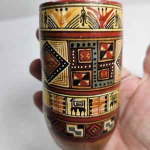 May include: A small, cylindrical ceramic cup with a brown base and intricate geometric patterns in red, yellow, black, and white.