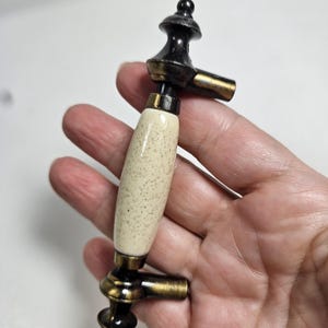 May include: A cabinet pull with a speckled cream-colored ceramic handle and bronze-toned metal accents. The pull has a decorative design with a black top and a curved shape, suitable for furniture restoration or new builds.