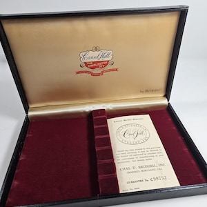 May include: An open black box with a burgundy velvet interior. The lid's interior is tan with the words "Carvel Hall The Charleston Set" in a decorative logo. A guarantee card is visible.