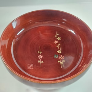 May include: A round, reddish-brown wooden bowl with a glossy finish. The interior features a floral design with pink, green, and gold accents. A small square stamp is visible near the floral design. The bowl's rim is a lighter shade of brown.