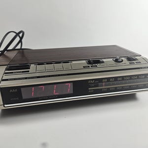 May include: A vintage GE electronic digital FM/AM clock radio. The clock has a silver and brown finish with a digital display showing the time. It features buttons for snooze, sleep, wake, and hour settings. The radio dial displays FM and AM frequencies.