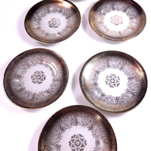 May include: A set of five round ceramic saucers featuring a white center with a central floral motif, surrounded by a wide, dark metallic border decorated with intricate, swirling filigree patterns in a reflective gold and black finish.