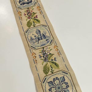 May include: A long, narrow, beige cross-stitch tapestry with three framed scenes of a sailboat, a lighthouse, and a building. The tapestry is adorned with floral and geometric patterns in blue, green, and brown, and has a decorative metal edge.