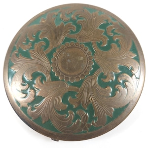 May include: A round metal compact case featuring a dark green enamel background with an embossed brass-toned floral scrollwork pattern. The center of the lid displays a circular medallion surrounded by a decorative beaded border design.