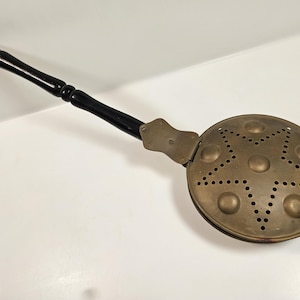 May include: Antique brass bed warmer with a long, black wooden handle. The round warming pan features a star-shaped pattern of perforations and decorative brass rivets. A vintage kitchenware item.