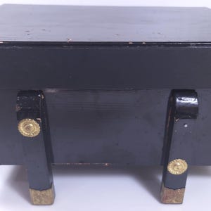 May include: A rectangular black box with a lid, featuring four vertical legs and decorative gold-toned metal corner accents. The legs are adorned with circular gold-toned metal hardware at the top and gold-toned metal caps at the base.