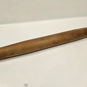 May include: A wooden rolling pin with a light brown finish. The rolling pin is cylindrical with a tapered end and is made of a light-colored wood. The wood grain is visible on the surface.