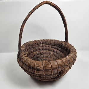 May include: A small, brown, woven basket with a single handle. The basket is made of natural materials and has a round shape.