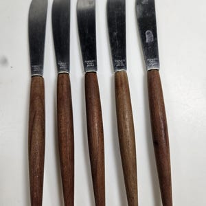 May include: Five butter knives with stainless steel blades and wooden handles. The blades are curved and the handles are a dark brown color. The knives are arranged in a row, with the blades facing up.