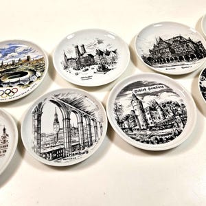 May include: A collection of seven white ceramic souvenir plates featuring black and white illustrations of German landmarks, including Munich, Hamburg, and Cologne, with one plate displaying a colorful view of the 1972 Olympic Park in Munich.
