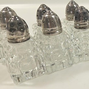 May include: Six clear glass salt and pepper shakers with silver-colored tops. The shakers have a square, textured design. The tops have small holes for dispensing the salt and pepper.