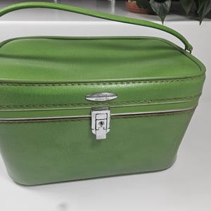 May include: Green vintage cosmetic case with a curved handle and silver-tone hardware. The case has a rectangular shape with rounded edges and a secure latch. The exterior is a solid, textured green with a stitched trim. The brand name "Samsonite" is visible.