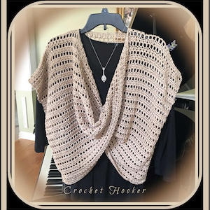 May include: A beige crochet top with a criss-cross design and a V-neck. The top is hanging on a black hanger and is on a white piano.