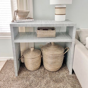 May include: A white side table with two shelves, a white lamp with a woven shade, a wooden bowl with a beaded garland, a book titled "Homebody", and two woven baskets.
