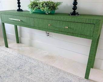 May include: A green console table with two drawers and clear acrylic handles. The table has a textured surface and angled legs. A green planter with greenery sits on top, with two black lamps on either side.