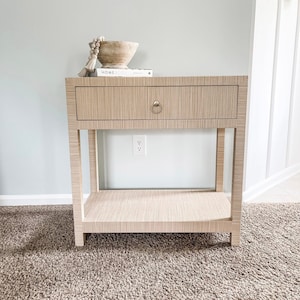 May include: A light wood end table with a single drawer and a lower shelf. The table has a simple, modern design and is perfect for a bedroom or living room. The drawer has a gold knob and the table is sitting on a beige carpet.
