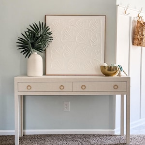 May include: A white console table with two drawers and gold ring pulls. The table is decorated with a white framed wall art with a raised design, a white vase with green leaves, and a gold bowl with decorative items.