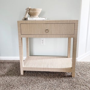 May include: A light wood end table with a single drawer and a lower shelf. The drawer has a brass knob. The table is sitting on a beige carpet. The text "HOME BODY" is visible on a book on top of the table.