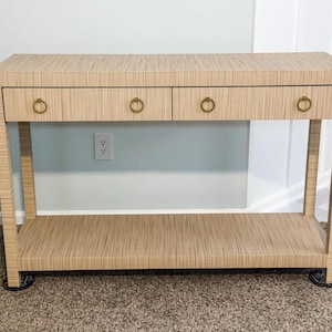 May include: A light beige console table with two drawers and a lower shelf. The drawers have brass ring pulls. The table has a textured, vertical striped pattern. The legs are straight and the table is on a carpeted floor.