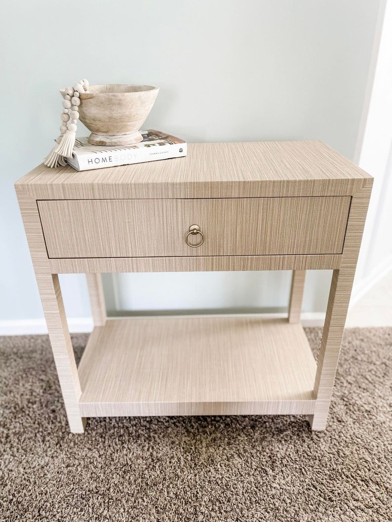 May include: A light beige nightstand with a single drawer and a lower shelf. The drawer has a gold knob. A book titled "Homebody" is on top of the nightstand, along with a wooden bowl and a white beaded garland.