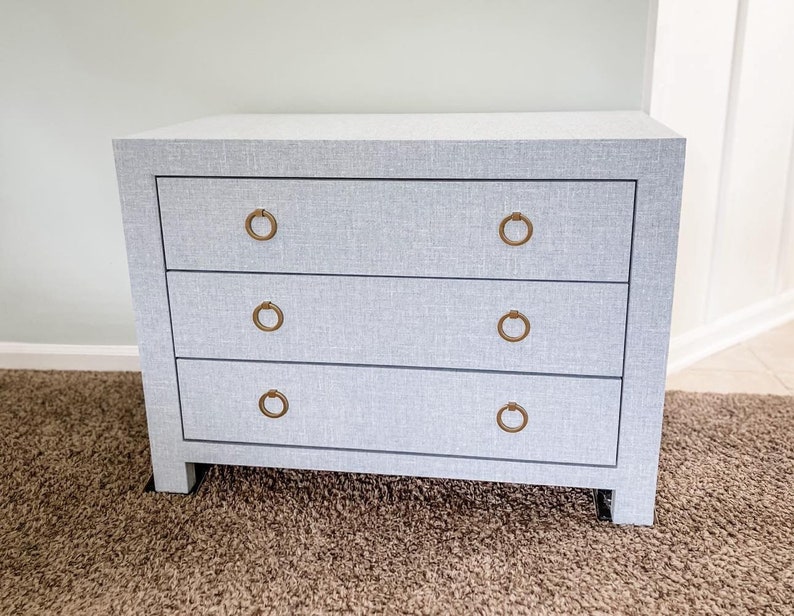 May include: A light gray dresser with three drawers and brass ring pulls. The dresser is sitting on a brown carpet.