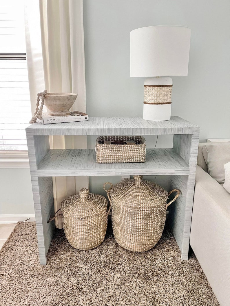 May include: A white side table with two shelves, a white lamp with a woven shade, a wooden bowl with a beaded garland, a book titled "Homebody", and two woven baskets.