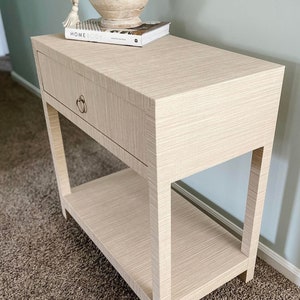 May include: A light beige end table with a single drawer and a lower shelf. The table has a woven texture and a simple, modern design. The table is topped with a book and a wooden bowl with white beads.