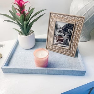 May include: A blue tray with a woven texture holds a pink candle, a white planter with a green plant with pink flowers, and a framed black and white photo.