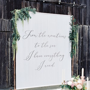 Puede incluir: Un telón de fondo blanco con el texto "From the mountains to the sea I have everything I need." en caligrafía negra. El telón de fondo está instalado en un soporte de cobre frente a una pared de madera. Una mesa con un mantel blanco y flores rosas está en primer plano.