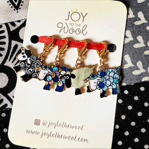 May include: Four colourful sheep stitch markers with gold clasps. The sheep are black and white, blue and white, and multi-coloured. The markers are on a white card with the text "Joy to the Wool" and a website address.