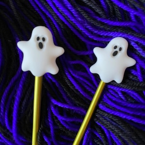 May include: Two gold knitting needles with white ghost-shaped tops. The needles are lying on a pile of black and purple yarn.