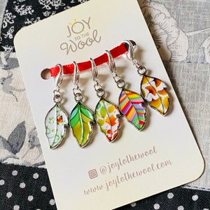 May include: Set of six colourful leaf-shaped stitch markers with silver metal rings. The markers are decorated with different patterns and colours. The markers are displayed on a white card with the text "Joy to the Wool" and a social media handle.
