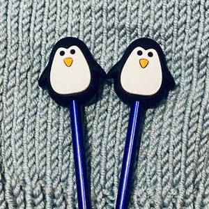 May include: Two blue knitting needles with penguin-shaped tops. The penguins are black and white with orange beaks.