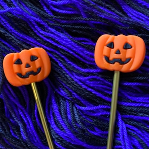 May include: Two orange pumpkin-shaped knitting needles with black eyes and a smiling mouth, set against a background of purple and black yarn.