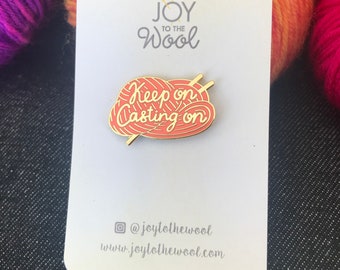 Könnte beinhalten: Eine goldene Emaille-Anstecknadel mit einer roten Wollknäuel und Stricknadeln. Die Nadel zeigt den Text "Keep on Casting on" und oben den Text "Joy to the Wool".